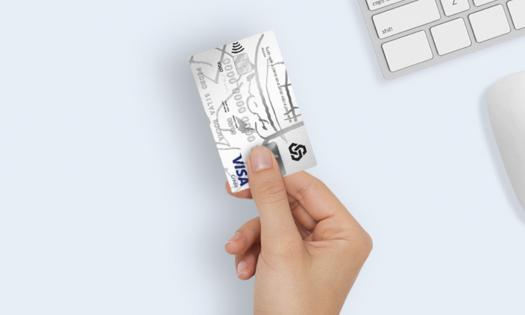 Mão segurando cartão de crédito Caixa In; detalhe de teclado e mouse