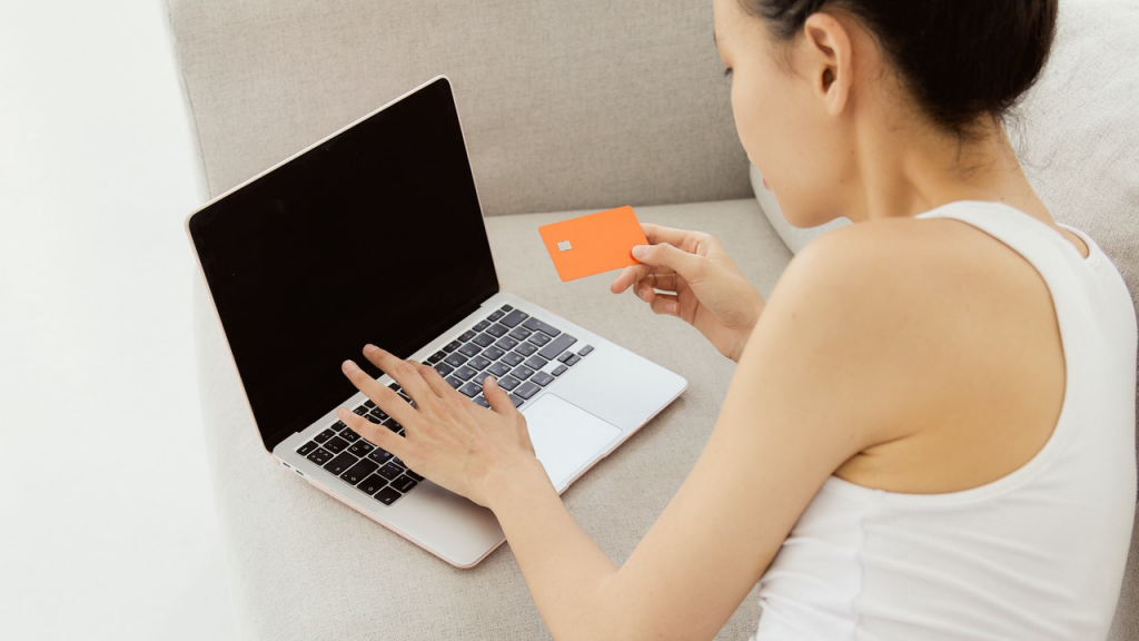 Mulher mexendo em notebook sobre sofá e segurando cartão laranja de débito BPI Electron