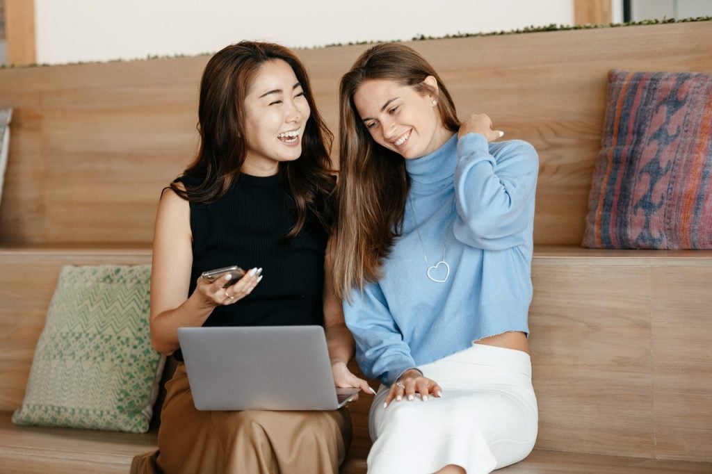 Duas mulheres sentadas conversando sorrindo, uma delas com notebook no colo e segurando celular