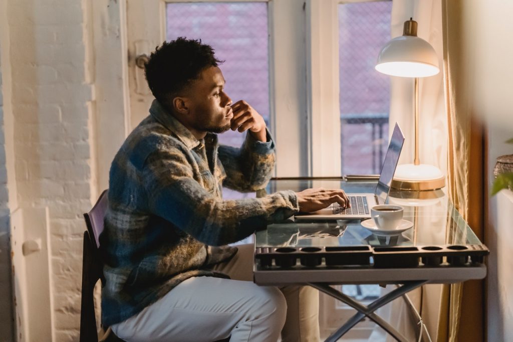 Homem em escritório em casa usando notebook. Luminária e xícara sobre a mesa.