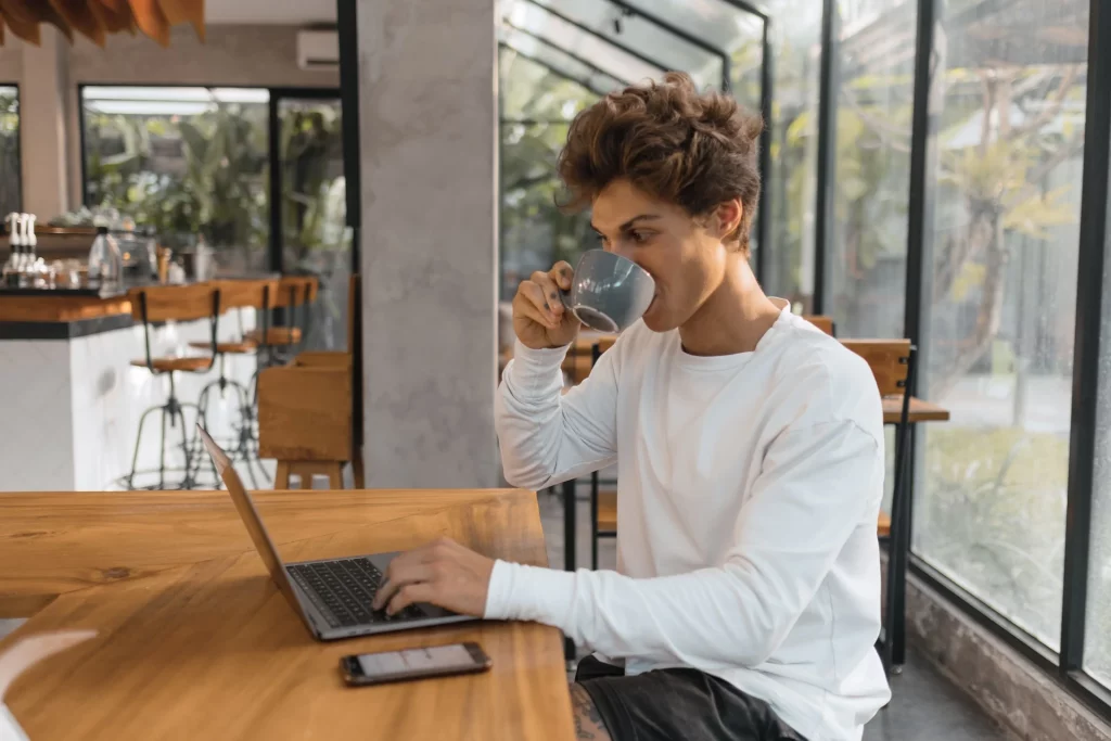 Pessoa bebendo de uma caneca/xícara enquanto navega online em notebook, celular ao lado de notebook. Ambiente moderno com janela para o ar livre com árvores desfocada ao fundo