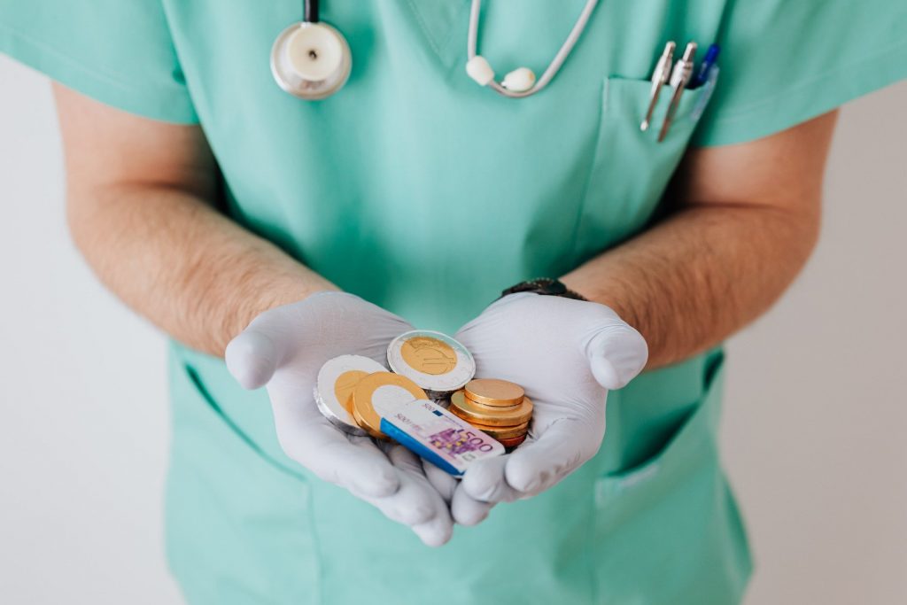 Pessoa com uniforme hospitalar, com estetoscópio pendurado nos ombros, usando luvas de latex segurando moedas e nota de euro