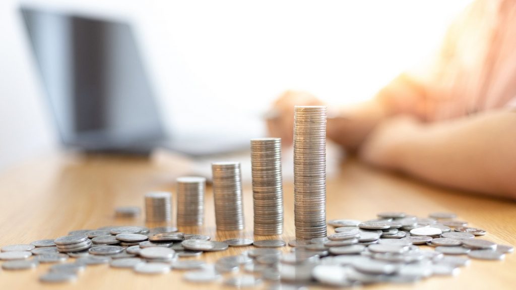 Trilha de moedas crescendo, representando investimento, com moedas ao redor sobre a mesa, pessoa mexendo em notebook no fundo desfocado