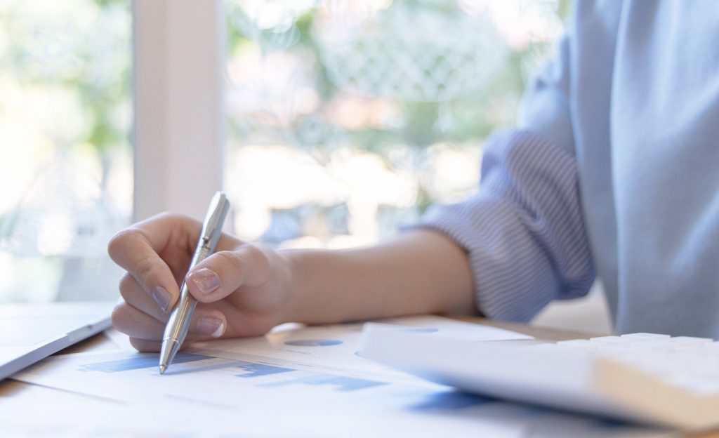 Pessoa com blusa azul (cor da identidade visual da corretora Degiro), com calculadora ao lado sobre a mesa, escrevendo com caneta metálica num papel com gráfico crescente, representando como funciona os investimentos na corretora