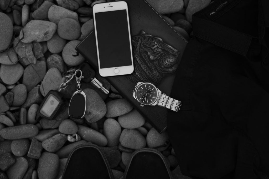 Celular (onde pode ser solicitado o crédito Creditas), relógio, chave de carro, caderno, camiseta e tênis sobre rochas