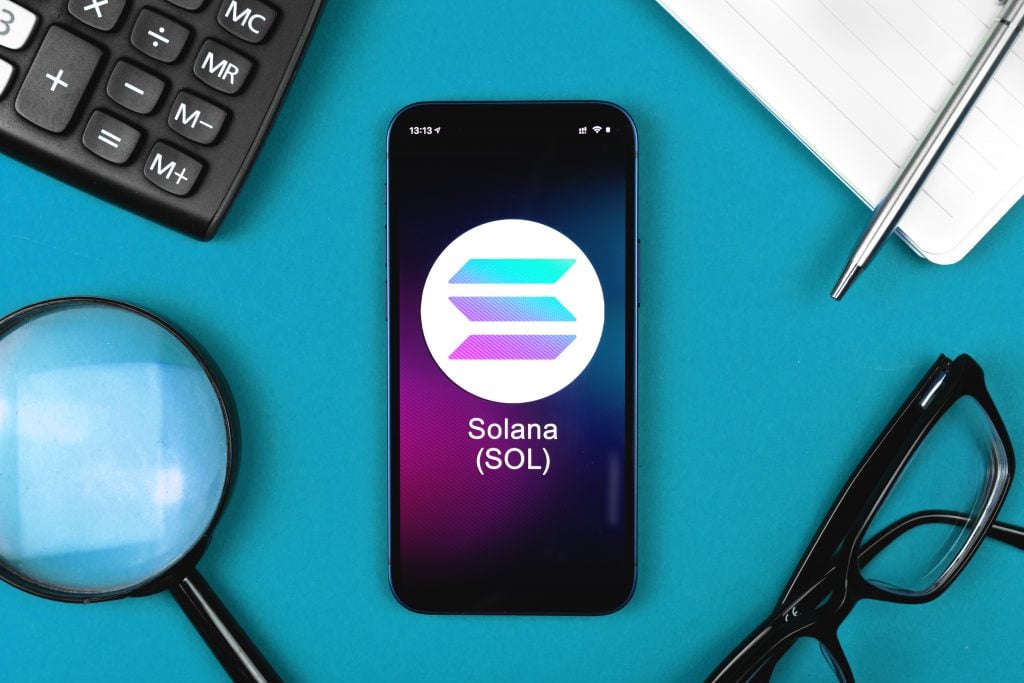 Logotipo da criptomoeda Solana, Solana e SOL escritos em tela de celular. Calculadora, lupa, óculos, papel e caneta ao redor do celular; sobre mesa azul.
