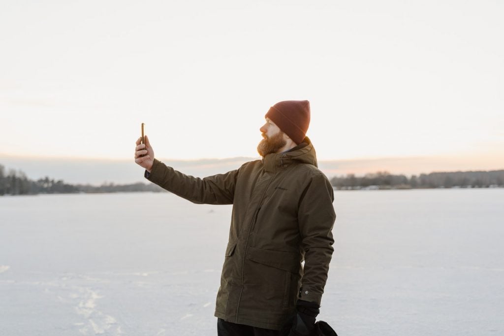 Homem ao ar livre, lago ao fundo, com touca e jaqueta, segurando celular - no qual é possível aderir à corretora AvaTrade