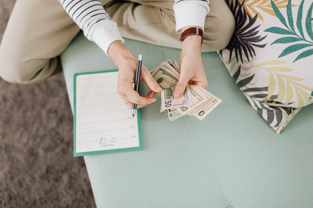 imagem com fundo claro de uma pessoa sentada contando notas de dinheiro e uma agenda com anotações