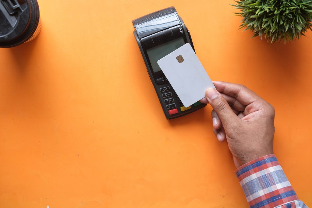 Superfície laranja e vasos de planta ao redor. Mão aproximando cartão contactless de maquininha de pagamento. Simbolizando o cartão de débito Novo Banco.