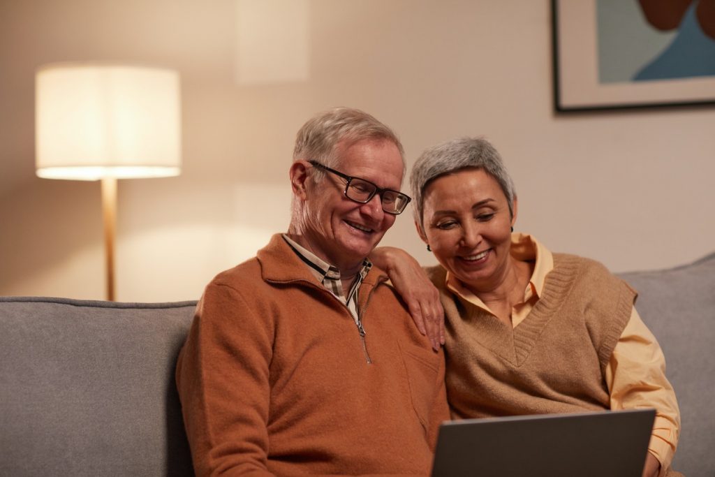 Casal de idosos em sala de estar, sentados no sofá, sorrindo e usando um notebook. Simbolizando como contratar o Crédito Montepio