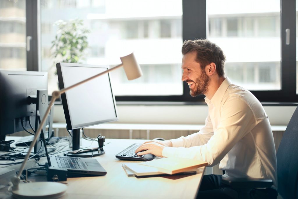 Homem de camisa branca social em escritório, sorrindo olhando para tela de computador enquanto digita no teclado, simbolizando como solicitar o crédito Cifra7 online
