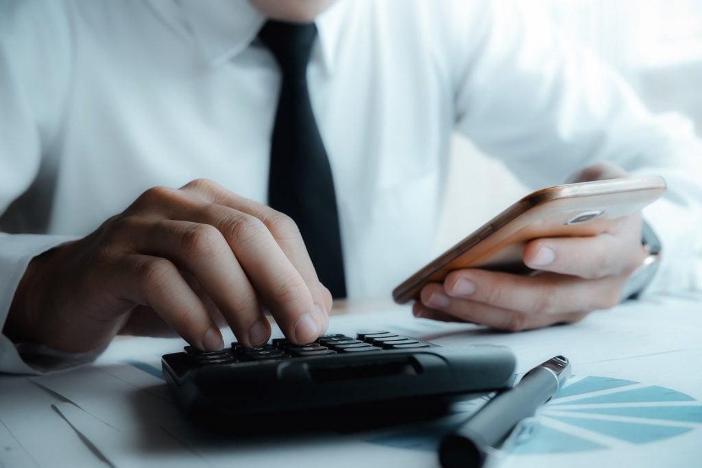 Homem de camisa branca e gravata preta segurando celular enquanto usa uma calculadora. Simbolizando e-Faturas