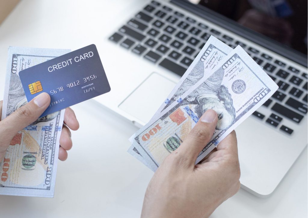 imagem com uma pessoa segurando um cartão de crédito, algumas notas de dinheiro em frente a um computador para pesquisar sobre obter bonificação no spread do crédito