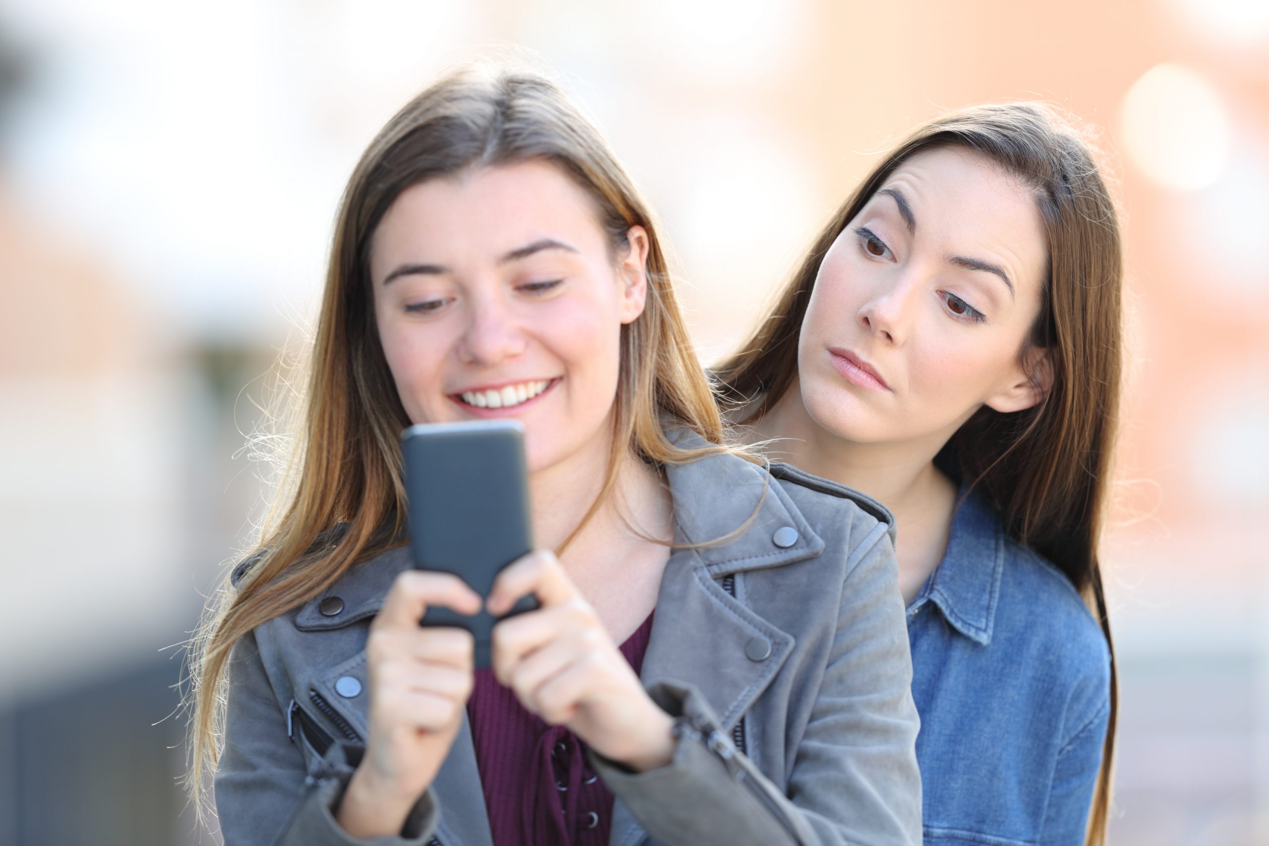 Mulher sorrindo usando celular e outra mulher desconfiada espiando a tela por cima do ombro