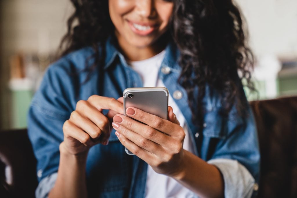 Cropped shot of an african-american young woman using smart phone at home. Smiling african american woman using smartphone at home, messaging or browsing social networks while relaxing on couch