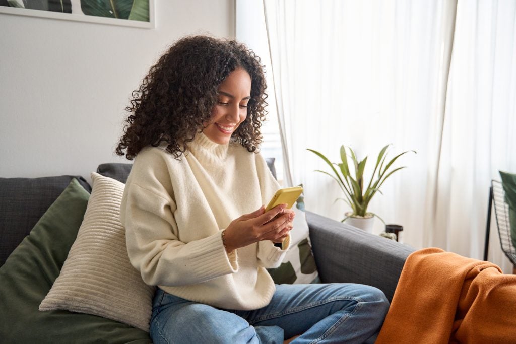 Happy young latin woman sitting on sofa using phone in living room.