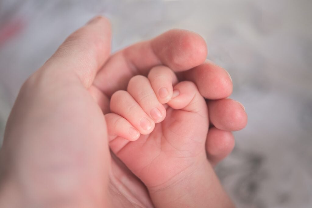 Mão de adulto envolvendo a mão de um bebê, simbolizando a importância de solicitar Auxílio Filho