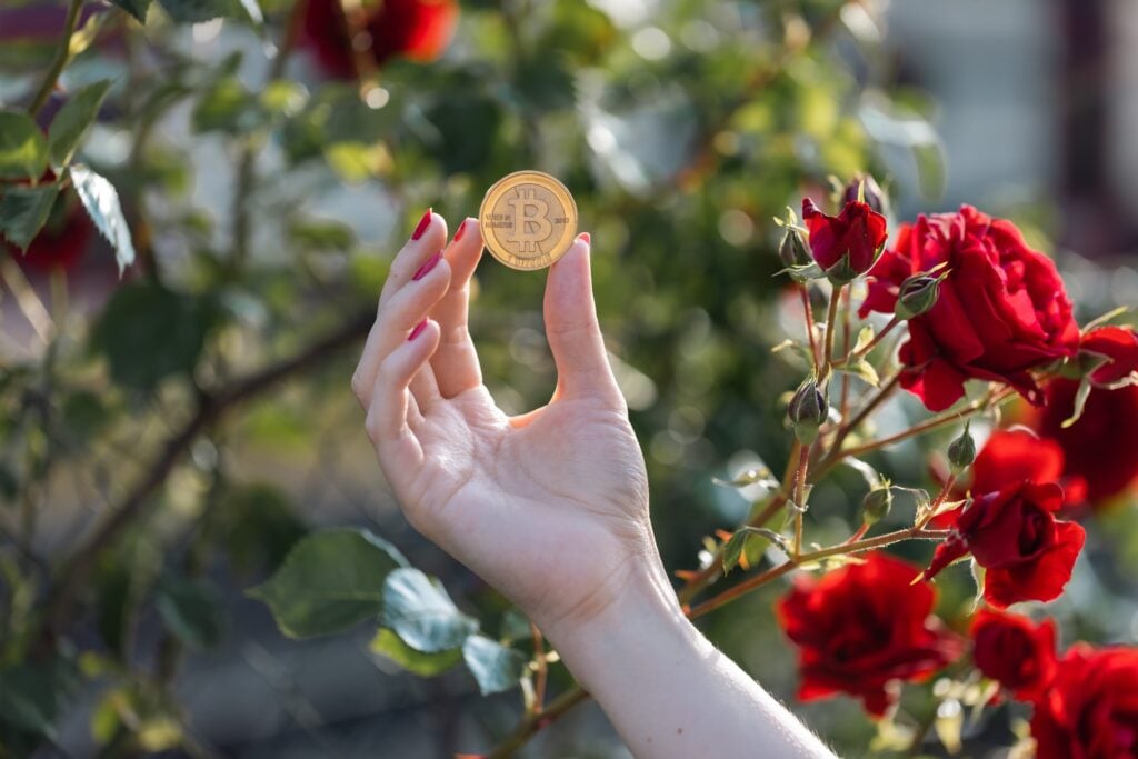 Mão com esmalte segurando moeda de Bitcoin, fundo com rosas vermelhas. Simbolizando investimento em Crypto FX com o Banco Carregosa