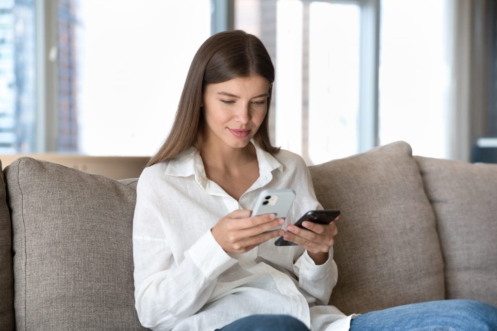 Mulher sorrindo sentada no sofá e olhando dois celulares
